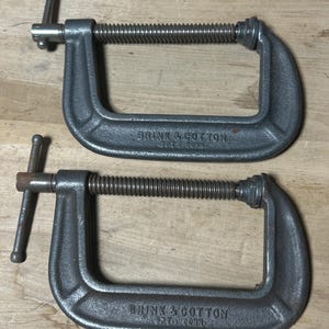 May include: Two gray C-clamps with threaded rods and handles. The clamps are marked "BRINK & COTTON" and are designed for woodworking or metalworking projects. The clamps are made of cast iron and are used to hold materials together.