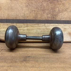 May include: A vintage metal door knob with a round, slightly domed shape. The knob is a dark gray color and has a smooth, slightly textured surface.