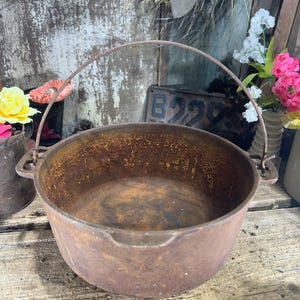 May include: A rusty, cast iron cauldron with a curved handle. The pot has a rounded shape and is weathered with signs of age. The interior shows signs of use. The cauldron is likely used for cooking or decorative purposes.