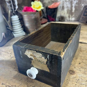 May include: A rectangular wooden drawer with a dark, weathered finish and a single white ceramic knob. A torn, aged paper label is partially attached to the front face, featuring faint, partially visible text that includes the letters ING.