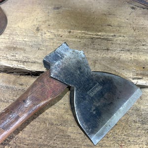 May include: A vintage hatchet with a weathered steel head and a dark brown wooden handle. The metal head features a distinct flared blade shape and a visible stamped logo on the side. The tool shows signs of age with surface oxidation.