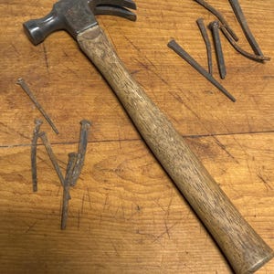 May include: A vintage claw hammer with a wooden handle and a set of rusty nails on a wooden surface.