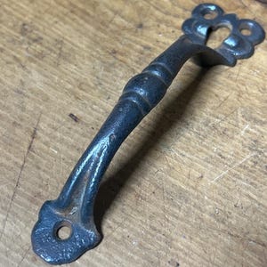 May include: A dark gray, ornate metal handle with a decorative floral design. The handle has a curved, ridged grip and is likely for a cabinet or drawer. The metal has a slightly aged, rustic appearance.