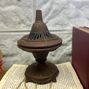 May include: A small, ornate, bronze incense burner with a decorative top and a base with intricate details. The burner is sitting on a stack of old books.