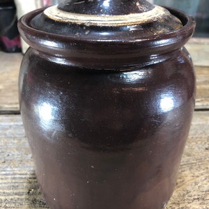 May include: A brown glazed ceramic crock with a lid. The lid has a knob on top and a white rim.