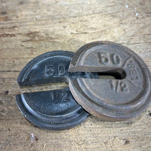 May include: Two circular cast iron slotted weights, each embossed with the text 50 and 1/2 LBS. The weights feature a dark, weathered metallic finish with visible signs of oxidation and wear, showing a distinct slot cut into the side.