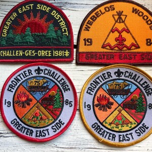 May include: Four embroidered patches with the text "Greater East Side" and various scout-related designs. The patches are in different colors, including red, orange, yellow, and blue. The patches feature a campfire, a teepee, a wagon, and a sunrise.