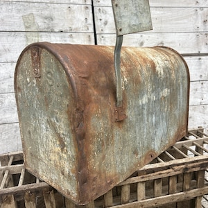 May include: Weathered metal mailbox with a curved top and a small flag. The mailbox has a rusty, aged appearance, with visible signs of wear and tear. The mailbox is sitting on a wooden crate.
