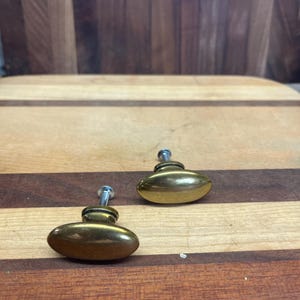 May include: Two oval-shaped brass cabinet knobs with a silver screw. The knobs are on a wooden surface.