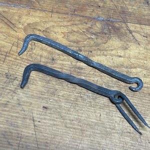 May include: Two hand-forged metal tools with a rustic appearance. One has a curved hook at the end, while the other features a looped handle and two prongs. The tools are displayed on a wooden surface, showcasing their craftsmanship.