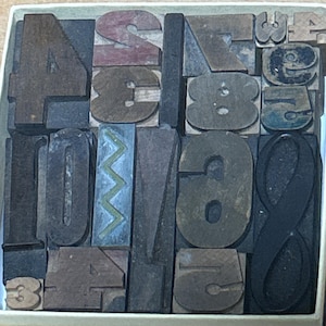 May include: A collection of vintage wooden letterpress blocks arranged in a square box. The set includes various letters, numbers, and symbols in different sizes, fonts, and shades of brown, black, and grey, with one block featuring a green zigzag.