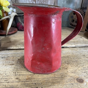 May include: A vintage red metal pitcher with a handle. The pitcher has a slightly worn appearance, with visible signs of age and use. The handle is curved and attached to the side of the pitcher. The pitcher is sitting on a wooden surface.