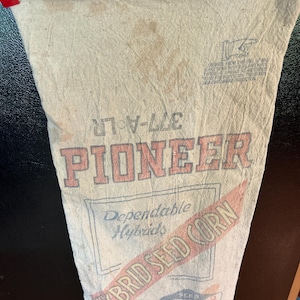 May include: A vintage beige fabric seed bag featuring the red text PIONEER and HYBRID SEED CORN. The bag includes the text 377-A-LR, Dependable Hybrids, and a small Pioneer Seed Corn logo. The material shows signs of age with light brown stains.