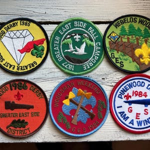 May include: A collection of six vintage embroidered patches. The patches feature various designs, including a diamond, a bridge, axes, and text such as "Pinewood Derby" and "Webelos Woods." Colors include yellow, green, red, and blue.