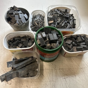 May include: A collection of metal letterpress type pieces stored in various containers, including a small metal bucket, several plastic food tubs, and a green plastic coffee canister, all filled with assorted rectangular lead type blocks.
