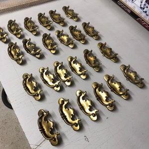 May include: A collection of 20 ornate brass drawer pulls with a decorative, scrolled design. The pulls are arranged in rows on a white surface.