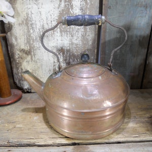 May include: A vintage copper kettle with a wooden handle and a black knob on the lid. The kettle has a worn, aged look and is sitting on a wooden surface.