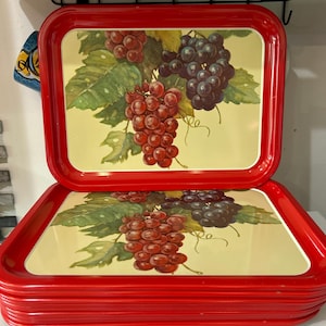 May include: A stack of red metal serving trays. Each tray features a painted design of red and purple grapes with green leaves on a yellow background. The trays are rectangular with rounded corners and a raised edge.