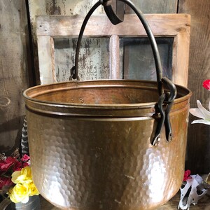 May include: A large, hammered copper pot with a hanging iron loop handle. The pot is empty and has a shiny, metallic finish.