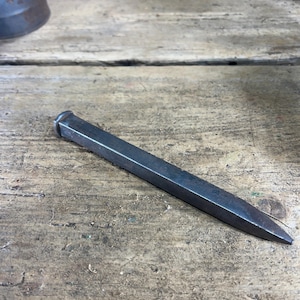 May include: A dark gray, hand-forged iron railroad spike with a square-shaped shank and a flat, rounded head. The metal surface shows a weathered, matte texture with subtle variations in tone, tapering to a sharp, pointed end at the base.