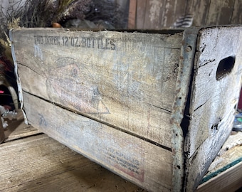 Könnte beinhalten: Eine antike Holzkiste mit Metallverzierungen. Die Kiste ist mit "Two Dozen 12 oz Bottles" beschriftet.