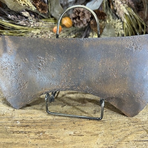 May include: A rusty, weathered, double-bit axe head with a rounded edge. The axe head is displayed on a black metal stand.