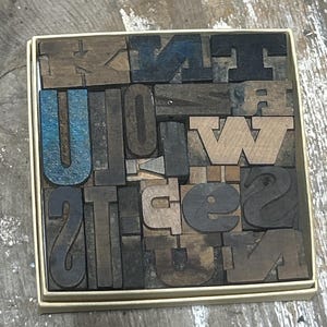 May include: A square box filled with an assortment of vintage wooden letterpress blocks in various sizes and fonts. The blocks are arranged in a grid, featuring a mix of natural wood tones, dark stained wood, and some pieces with blue paint.