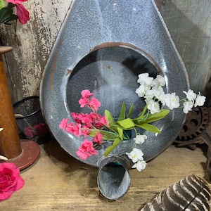 May include: A vintage, speckled gray enamelware chamber pot with a rounded opening and a spout. The pot is decorated with pink and white flowers. A large feather and other objects are in the background.