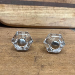 May include: Two clear glass hexagonal knobs with a silver center. The knobs are on a wooden surface.