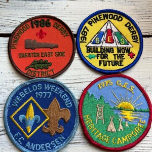May include: Four round embroidered patches with a scout theme. The top left patch is red with the text "Pinewood 1986 Derby" and "Greater East Side District". The top right patch is yellow with the text "1987 Pinewood Derby" and "Building Now For The Future". The bottom left patch is blue with the text "Webelos Weekend 1977" and "E.C. Andersen". The bottom right patch is blue with the text "1975 G.E.S. Heritage Camporee".