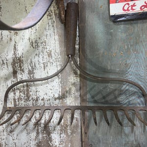 May include: A vintage garden rake with a wooden handle and curved metal tines. The rake is made of weathered metal, showing signs of age and use. The handle is a dark brown color. The background is a weathered wooden wall.