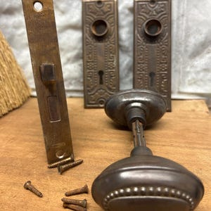 May include: Antique brass door hardware set with a  knob, two plates, and screws. The plates have an ornate design and the knob has a beaded edge.