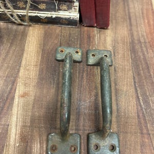 May include: Two vintage metal handles with a weathered, aged appearance. Each handle has a rectangular base with two holes for mounting and a curved, arched grip. The handles are a muted, greenish-gray color.