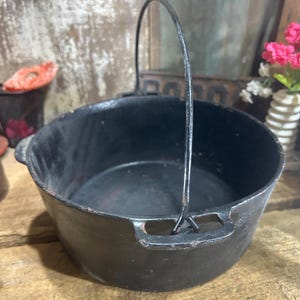 May include: A round, black cast iron pot featuring a deep basin, two side handles, and a tall, arched metal bail handle. The exterior and interior surfaces show a dark, matte finish with subtle signs of wear and slight rust along the edges.