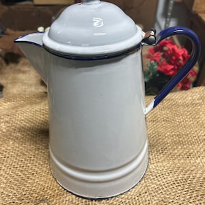 May include: A white enamel coffee pot featuring a dark blue rim, a matching dark blue handle, and a dark blue knob on the lid. The pot has a tapered cylindrical body with a prominent spout on the left side and a hinged lid mechanism at the top.