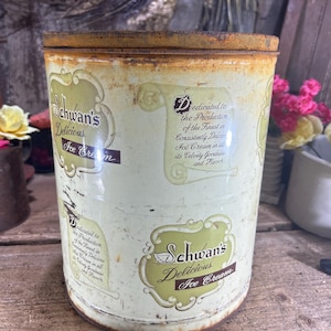 May include: Vintage Schwan's ice cream container. The cylindrical metal container has a rusty lid and a cream-colored body. The container features the text "Schwan's Delicious Ice Cream" in a green and brown design. The container also has text that reads "Dedicated to the Production of the Finest in Consistently Delicious Ice Cream."
