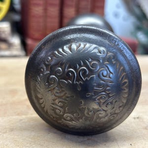 May include: A round, dark metal door knob featuring an intricate, embossed floral and scrollwork pattern across its surface. The knob has a weathered, aged finish with visible texture and depth, showcasing a detailed relief design on the front face.