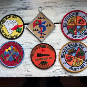 May include: Six embroidered patches with various designs, including a diamond, a scout emblem, a woodsman, a car, and a frontier challenge. The patches are all circular and have a red, white, and blue color scheme.