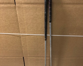 May include: Two golf clubs with black grips and silver shafts. The clubs are leaning against a brown cardboard box.