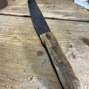May include: A vintage-style butcher knife with a dark blade and a wooden handle. The handle is secured with three rivets. The knife is set against a rustic wooden surface, suggesting a focus on kitchen or culinary tools.