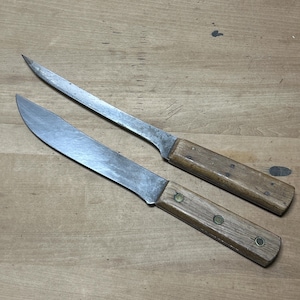 May include: Two vintage kitchen knives with wooden handles and metal blades. The knives are on a wooden surface.