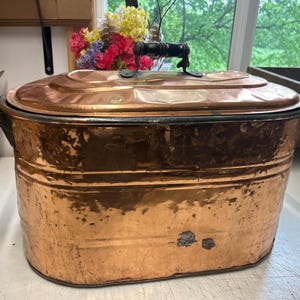 May include: A large, antique copper cooking pot with a lid and ornate handles. The pot has a rectangular shape with rounded edges and a shiny, aged finish. The lid features a black handle. A decorative item for the kitchen.