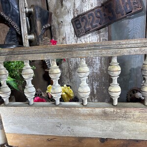 May include: A weathered wooden railing with five turned wooden balusters. The balusters are painted white with a distressed finish. The railing is mounted on a wooden wall with a license plate and a pair of old roller skates.
