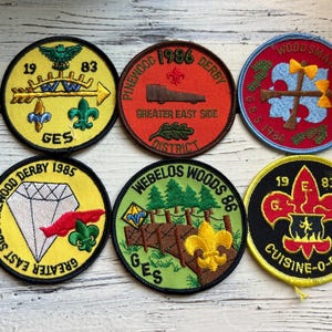 May include: Six colorful embroidered patches with the text "Pinewood Derby", "Webelos Woods", "Wood Sman Weekend", "Cuisine-O-Ree", and "GES" along with various designs including a diamond, a bridge, an axe, a cauldron, and an arrow.