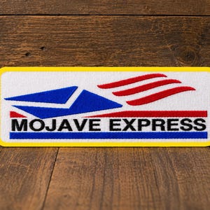 May include: A rectangular embroidered patch with a white background and a yellow border. It features a blue envelope icon, three red swooping lines, and the black text MOJAVE EXPRESS positioned above a thin blue horizontal line.