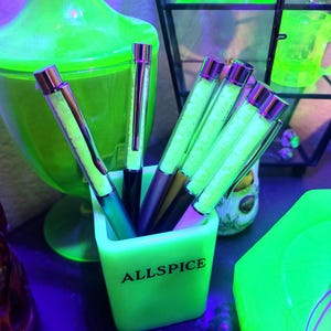 May include: A collection of pens in a neon green square container with the word "ALLSPICE" printed on the front. The pens have silver and purple accents. Other neon green objects are in the background.