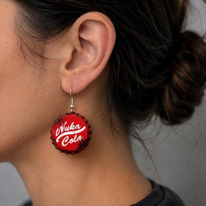May include: A dangling earring featuring a red bottle cap with a distressed, weathered texture. The text Nuka Cola is printed in white script across the center of the cap. The earring is attached to a silver-toned metal hook for ear piercing.