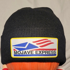 May include: A black knitted beanie hat with a rectangular patch displaying the words "MOJAVE EXPRESS". The patch has a blue and red design with a yellow border. The hat is made of a ribbed knit material.
