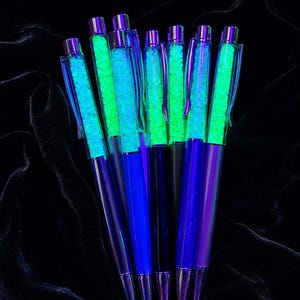 Pode incluir: Uma coleção de sete canetas com corpos metálicos em tons de azul e roxo. Cada caneta possui uma câmara superior transparente preenchida com pequenas partículas verde-neon brilhantes que se destacam no fundo escuro, com um clipe metálico.