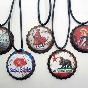 May include: Five bottle cap necklaces with different designs. The designs include a bottle of soda with a smiling face, a bear with a red star, a woman in a spacesuit, a bottle of soda with a red and white label, and a bottle cap with the text "Please Stand By".